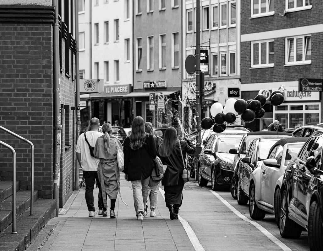 Streetphotography aus Köln: Freundesgruppe schlendert durch enge Altstadtgasse mit Ballons, Autos und Fassaden.