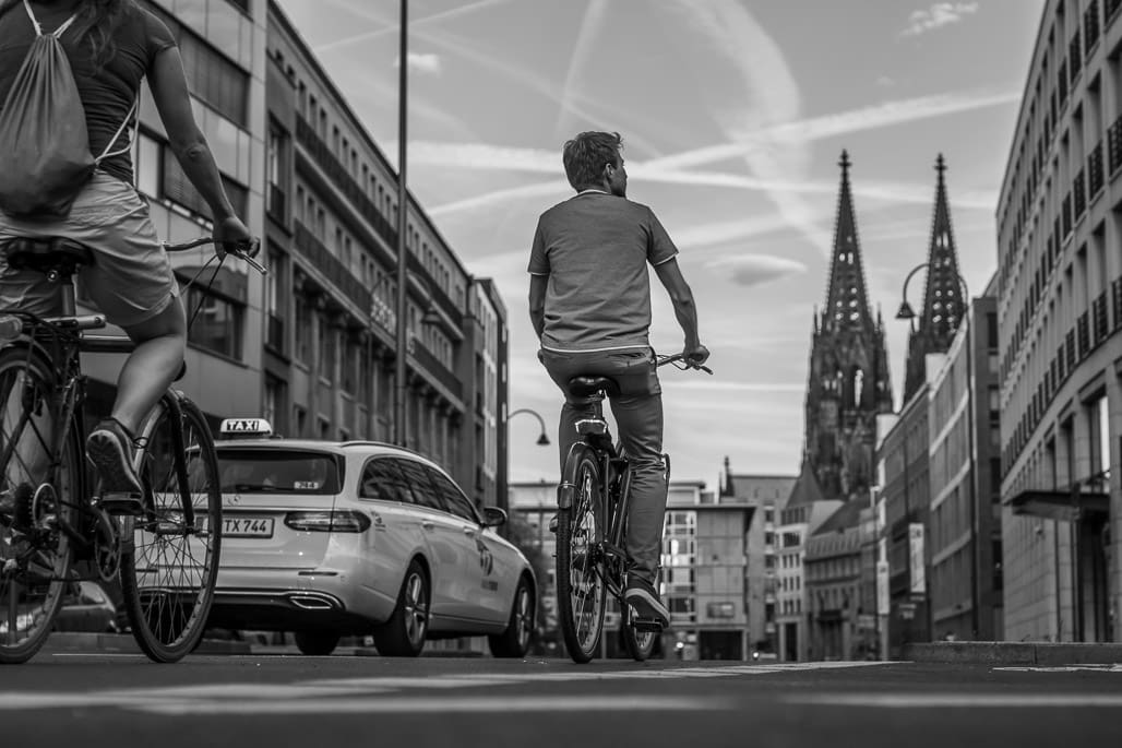 Streetphotography fängt Radfahrer vor Kölner Domkulisse und dramatischem Himmel mit Kondensstreifen ein.