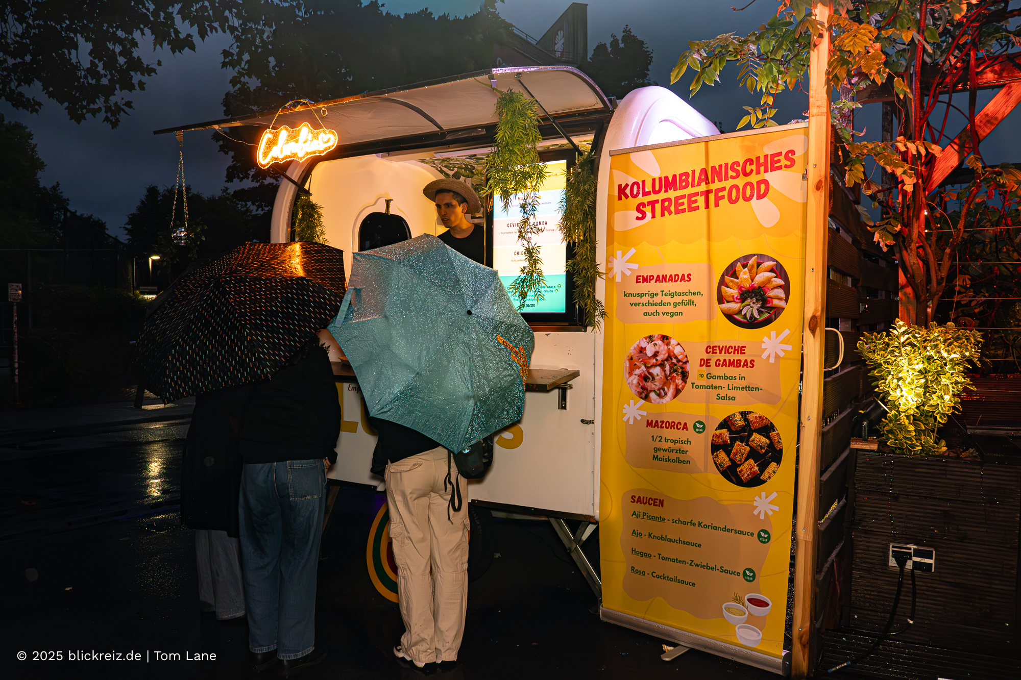 Streetfood-Stand mit kolumbianischen Spezialitäten auf der Hormiga Brava Messe Köln, Besucher mit Regenschirmen vor dem Foodtruck.
