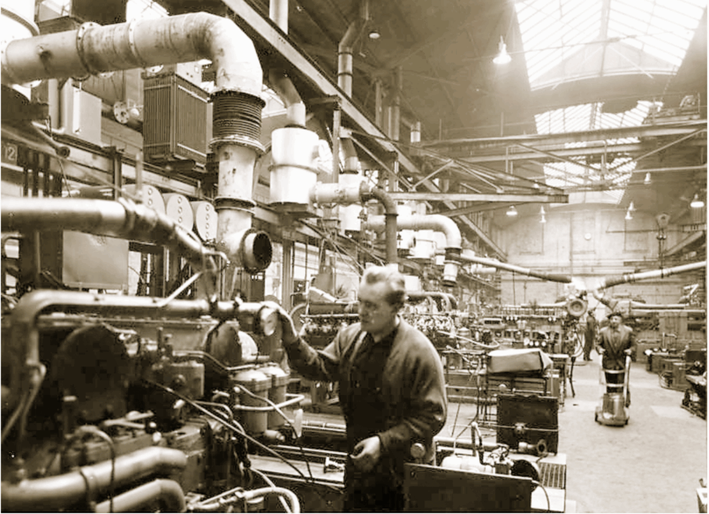 Historische Aufnahme aus den Hallen Kalk in Köln: Arbeiter bedienen schwere Maschinen in einer weitläufigen Industriehalle mit Rohrsystemen.