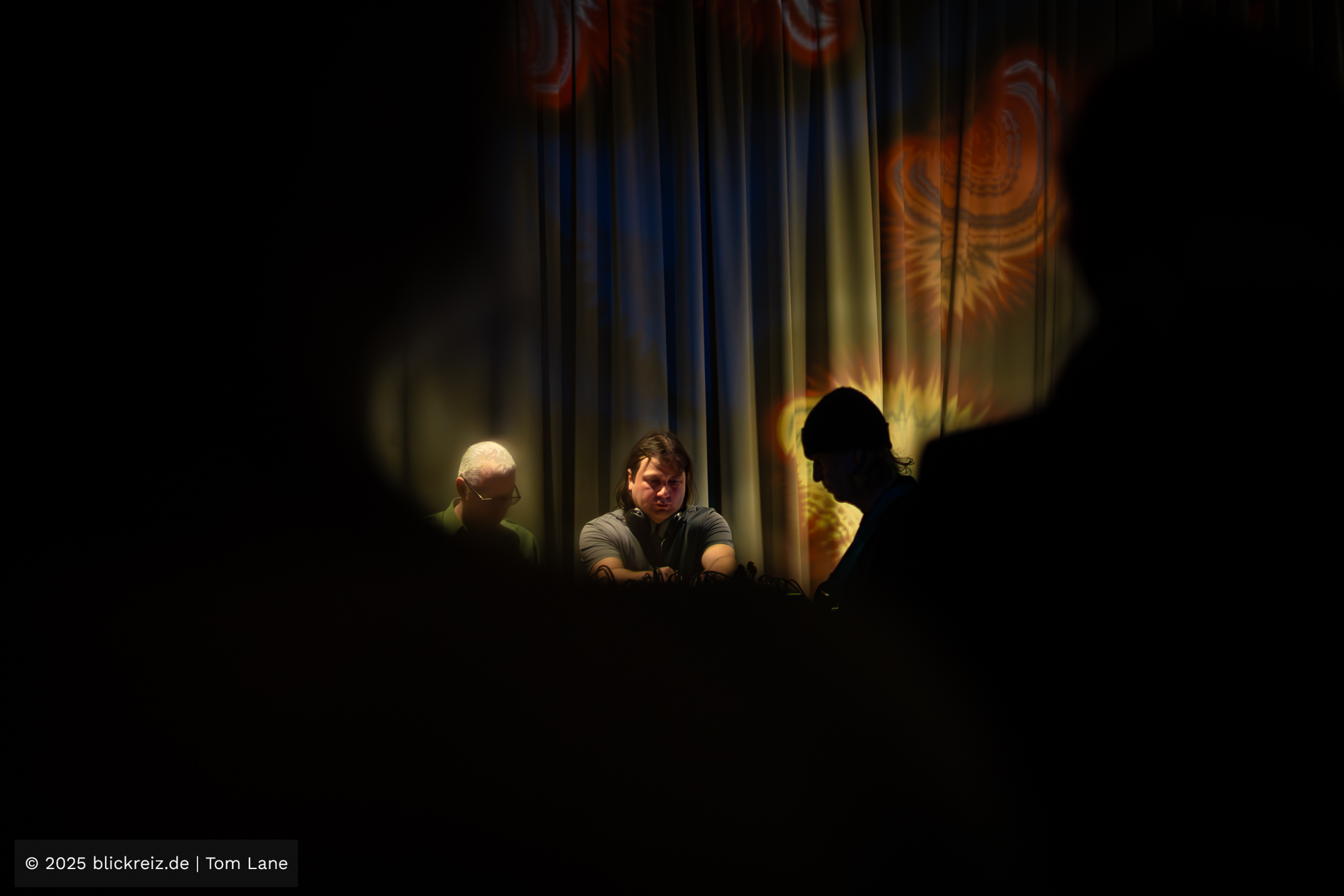 Ümit Han steht zwischen dunklen Silhouetten an seinem Setup, während farbige Projektionen den Hintergrund füllen und eine stille, konzentrierte Atmosphäre erzeugen.